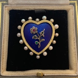 Może przedstawiać: Złota broszka w kształcie serca z niebieską emalią i kwiatowym wzorem. Broszka jest otoczona obramowaniem z pereł.