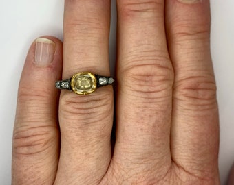 May include: A gold ring with a yellow gemstone surrounded by smaller diamonds. The ring is on a person's finger.