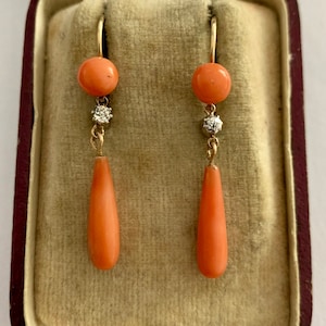 Pu&ograve; includere: Un paio di orecchini pendenti in corallo e diamanti in una scatola foderata in velluto. Gli orecchini sono in oro e presentano una perla di corallo rotonda e un ciondolo di corallo a forma di lacrima.