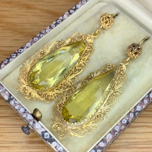 Op de afbeelding: Een paar gouden filigraan oorbellen met druppelvormige gele edelstenen. De oorbellen zitten in een klein wit doosje met een bruine, gepatenteerde voering.