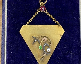 Può includere: Un ciondolo d'oro a forma triangolare con un design floreale che presenta una pietra preziosa verde, un diamante bianco e smalto nero. Il ciondolo è attaccato a una catena d'oro con una pietra preziosa rossa nella parte superiore.