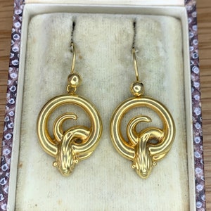 Op de afbeelding: Een paar gouden oorhangers met een cirkelvormig ontwerp met een knoop en een gestileerd dierenhoofd aan de onderkant.