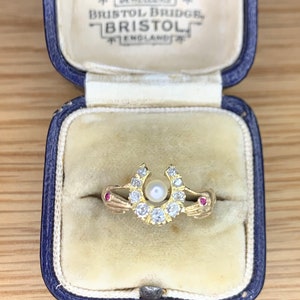 Puede incluir: Un anillo de oro con un diseño de herradura engastado con diamantes y una perla. El anillo está en una caja de anillo de terciopelo azul con la inscripción "FEARS LTD. WHOLESALE & EXPORT JEWELLERS BRISTOL BRIDGE, BRISTOL ENGLAND."