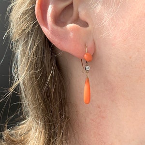 Pu&ograve; includere: Un paio di orecchini pendenti in corallo e diamante. Gli orecchini sono in oro e presentano un piccolo bead di corallo in cima, seguito da un piccolo diamante e poi da una goccia di corallo pi&ugrave; grande.
