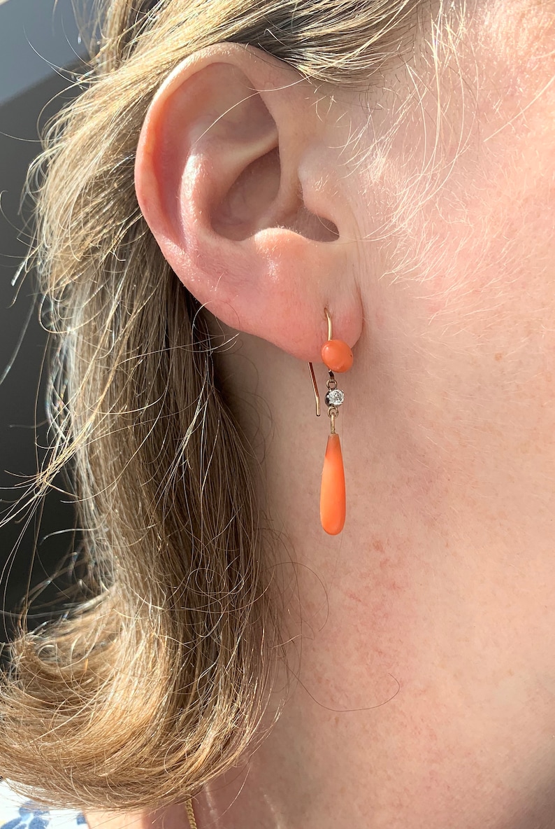 Pu&ograve; includere: Un paio di orecchini pendenti in corallo e diamante. Gli orecchini sono in oro e presentano un piccolo bead di corallo in cima, seguito da un piccolo diamante e poi da una goccia di corallo pi&ugrave; grande.