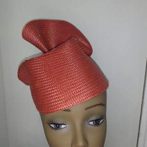 May include: A coral-colored hat with a woven texture, featuring a curved design. The hat is displayed on a mannequin head. The hat's design includes a folded, upward-sweeping brim.