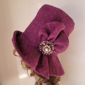 May include: A deep purple hat with a textured fabric and a large, decorative bow. The bow is adorned with a sparkling, multi-colored brooch. The hat has a wide brim and a structured shape, suitable for formal occasions.