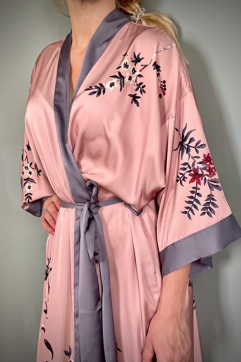 May include: A pink satin robe with a floral print and gray trim. The robe has a tie belt and long sleeves.