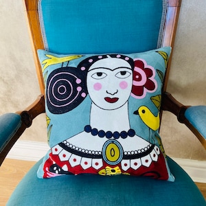 May include: A turquoise blue armchair with a decorative wooden frame and a colourful pillow featuring a stylised portrait of a woman with a red flower in her hair. The pillow has a blue background with yellow birds and black and white swirls.