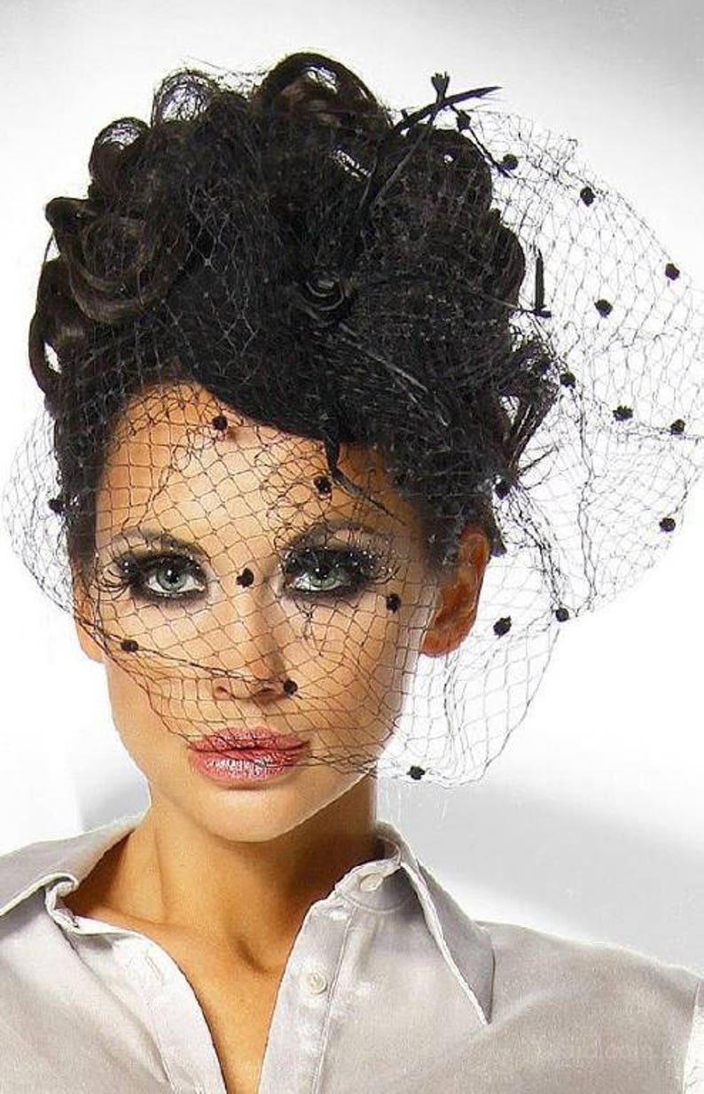 May include: A woman with dark hair styled in a bouffant wears a black veil with polka dots over her face. She is wearing a white collared shirt.