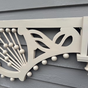 May include: Cream-colored wooden architectural detail with decorative cutouts and ball accents. The design includes a fan-like pattern and a small shelf with vertical slats. The piece is mounted on a gray surface.