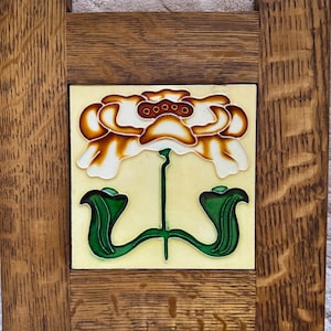 May include: A square ceramic tile featuring a raised, stylized floral design in shades of orange, white, and green, set within a dark brown, textured wooden frame with visible grain patterns and vertical side supports.