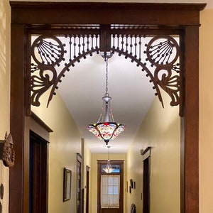May include: A hallway with a stained glass chandelier hanging from a decorative wooden archway. The archway has intricate carvings and a dark wood finish. The hallway has yellow walls and a light wood floor.
