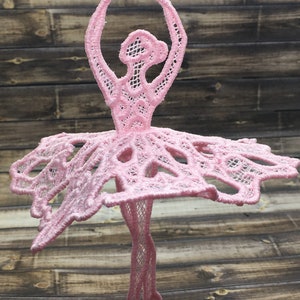 Könnte beinhalten: Ein rosa Ballerina-Anhänger aus Spitze mit einem filigranen Design. Die Ballerina befindet sich in einer Tanzpose mit erhobenen Armen über dem Kopf.