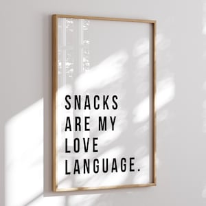 May include: A framed print with the text "SNACKS ARE MY LOVE LANGUAGE" in bold black letters on a white background. The frame is a light brown color, and the print is displayed on a white wall.
