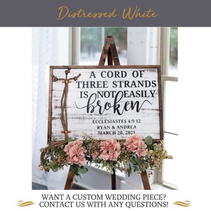 May include: A white wooden sign with distressed paint on an easel with pink flowers. The sign reads "A cord of three strands is not easily broken" with the scripture reference "Ecclesiastes 4:9-12" and the names "Ryan & Andrea" and the date "March 20, 2021".