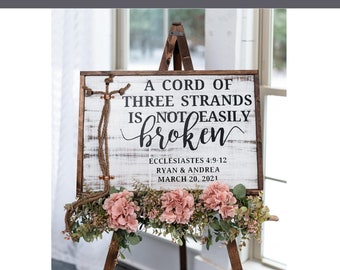 May include: A white wooden sign with distressed paint on an easel with pink flowers. The sign reads "A cord of three strands is not easily broken" with the scripture reference "Ecclesiastes 4:9-12" and the names "Ryan & Andrea" and the date "March 20, 2021".