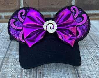 May include: A black baseball cap with purple and gold sequined ears and a large purple bow with a gold swirl embellishment.