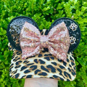 May include: A leopard print visor with black sequined mouse ears and a pink sequined bow. The ears have brown paw print embellishments.