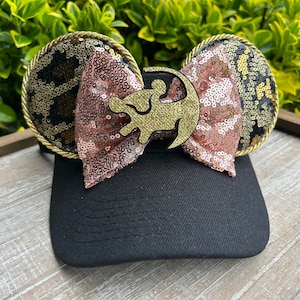 May include: Black baseball cap with a gold and brown leopard print bow and a gold lion silhouette. The bow is made of pink and gold sequins.