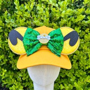 May include: Yellow baseball cap with attached mouse ears. The ears are yellow with black accents and gold trim. A green sequined bow with a gold center and a small, light blue embellishment is attached to the front of the cap.