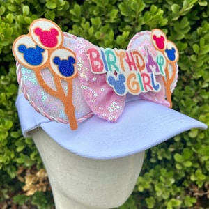May include: White visor with a pink sequined bow featuring the text "Birthday Girl". The bow is decorated with Mickey Mouse-shaped balloons in red, blue, and orange. A fun accessory for a birthday celebration.