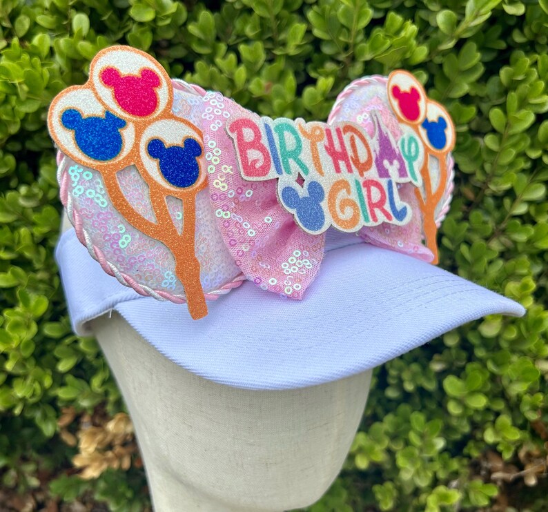 May include: White visor with a pink sequined bow featuring the text "Birthday Girl". The bow is decorated with Mickey Mouse-shaped balloons in red, blue, and orange. A fun accessory for a birthday celebration.