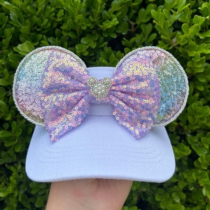 May include: A white visor with a large bow and two circular ears. The bow and ears are covered in iridescent sequins that shift colors. A small Mickey Mouse head is in the center of the bow, and the ears are trimmed with silver rope.