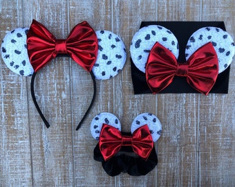 May include: Three sets of Dalmatian-print mouse ears with red bows. The largest set is on a headband, the medium set is on a headband with a black elastic band, and the smallest set is on a black elastic band.