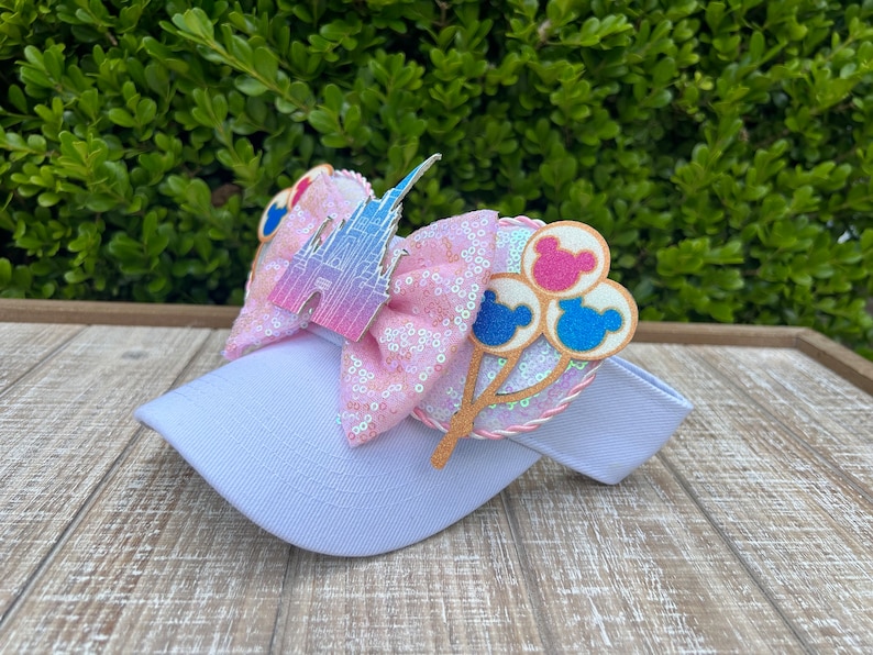 May include: A white visor with a pink sequined bow featuring a castle and Mickey Mouse balloon accents. The bow has a pink and white braided trim. The visor is on a wooden surface with green foliage in the background.