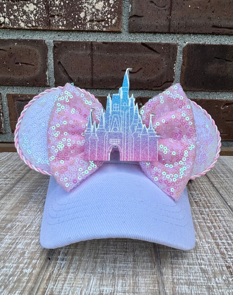 May include: A light purple baseball cap with a large pink sequined bow and a castle-shaped embellishment. The bow has a white and pink rope trim. The castle is a gradient of pink and blue.
