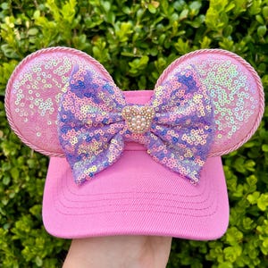 May include: Pink baseball cap with sequined mouse ears and a sequined bow. The ears are outlined with a pink rope trim. The bow features a gold-colored mouse head with pearl accents.