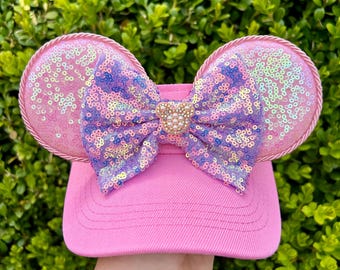 May include: Pink baseball cap with sequined mouse ears and a sequined bow. The ears are outlined with a pink rope trim. The bow features a gold-colored mouse head with pearl accents.