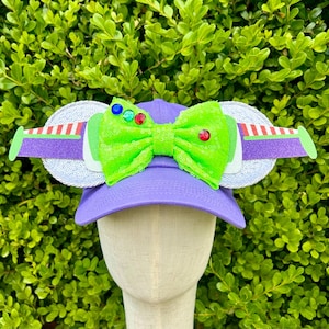 May include: Purple baseball cap with a green bow and white ears with a purple and white design. The ears have a red and white stripe and a green, blue, and red gem.