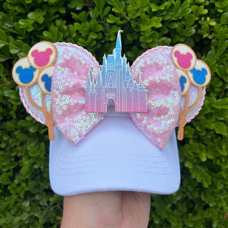 May include: White baseball cap with a pink sequined bow, featuring a castle and Mickey Mouse balloon accents. The bow is embellished with iridescent sequins. The castle and balloons are in shades of pink, blue, and gold.