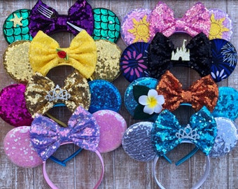Peut inclure: Une collection de bandeaux à oreilles de souris à paillettes de différentes couleurs, dont le jaune, le violet et le bleu. Chaque bandeau est orné d'un nœud décoratif et d'ornements thématiques tels qu'un diadème, une fleur ou un château. Les oreilles sont rondes et recouvertes de paillettes.