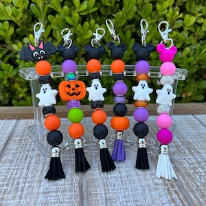 May include: Six Halloween-themed keychains with different charms, including a bat, pumpkin, ghost, and Mickey Mouse ears. Each keychain features colorful silicone beads and a tassel.