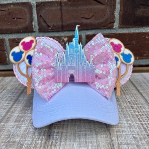 May include: A lavender baseball cap with a sequined pink bow and a castle-shaped embellishment. The cap features Mickey Mouse ear accents with balloon designs in pink, blue, and white. The ears are trimmed with a pink rope.