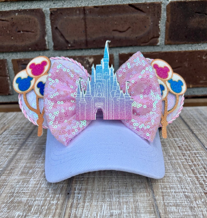 May include: A lavender baseball cap with a sequined pink bow and a castle-shaped embellishment. The cap features Mickey Mouse ear accents with balloon designs in pink, blue, and white. The ears are trimmed with a pink rope.