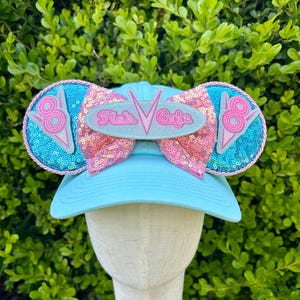 May include: A light blue baseball cap with a pink and blue bow and a "Twixt's Cafe" patch. The bow is made of sequins and the patch is a retro-style design.