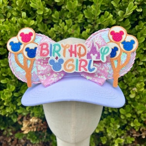 May include: A light purple baseball cap with a decorative bow and mouse ears. The bow is pink with sequins and the text "BIRTHDAY GIRL". The ears are decorated with mouse-shaped balloons in pink, blue, and red.