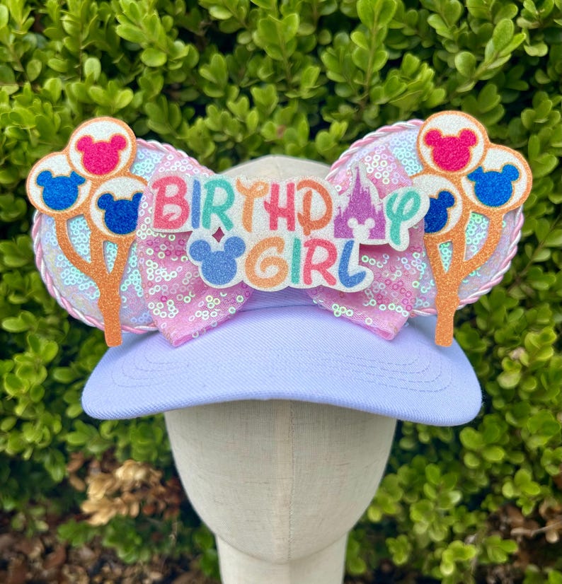 May include: A light purple baseball cap with a decorative bow and mouse ears. The bow is pink with sequins and the text "BIRTHDAY GIRL". The ears are decorated with mouse-shaped balloons in pink, blue, and red.