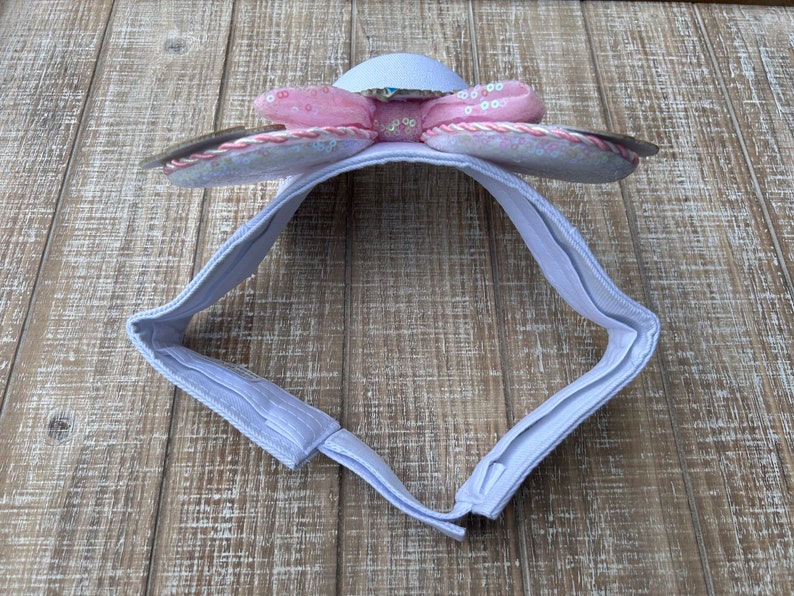 May include: A light purple headband with a flower design. The flower has pink sequined petals and a silver center. The headband is made of a soft fabric and has an adjustable closure.