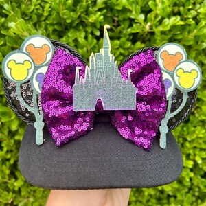 May include: Black baseball cap with purple sequin Minnie Mouse ears featuring a silver glitter castle and balloons with Mickey Mouse heads.