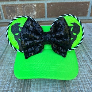 May include: A neon green baseball cap with black sequin bow and matching ear accents. The ears have a lime green background with a black silhouette design and are trimmed with white and black rope.