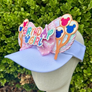 May include: A light blue visor with a pink sequined bow and decorative accents. The bow features the text "BIRTHDAY GIRL" and Mickey Mouse-themed balloon designs in pink, blue, and red. The visor is a fun accessory.