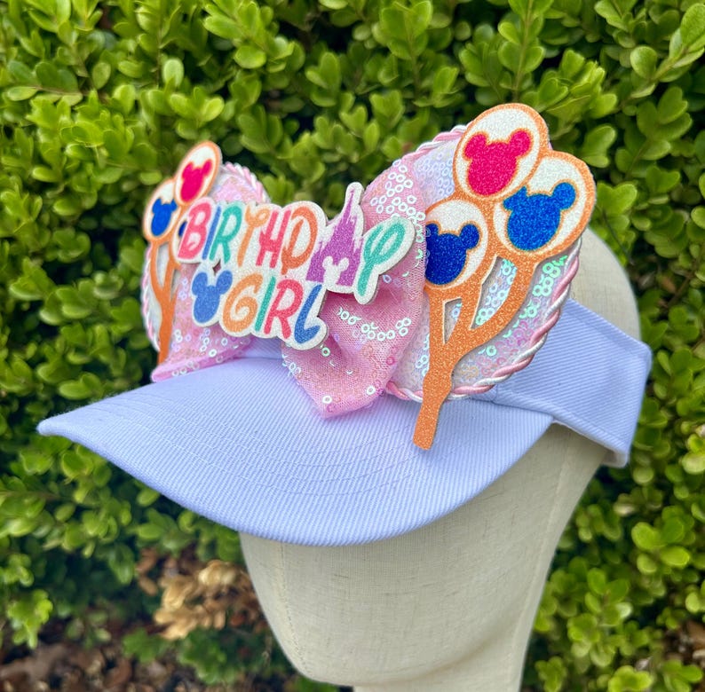 May include: A light blue visor with a pink sequined bow and decorative accents. The bow features the text "BIRTHDAY GIRL" and Mickey Mouse-themed balloon designs in pink, blue, and red. The visor is a fun accessory.