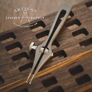 May include: A silver-toned leather crafting tool with a knob and two pointed ends, resting on a wooden surface with a grid pattern. The tool is labeled "Artisan Leather & Supply" with a compass logo.