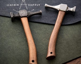 May include: Two metal hammers with wooden handles. The hammer heads are silver and dark gray, with a curved shape. The handles are a light brown color. The text "ARTISAN LEATHER SUPPLY" is at the top of the image.