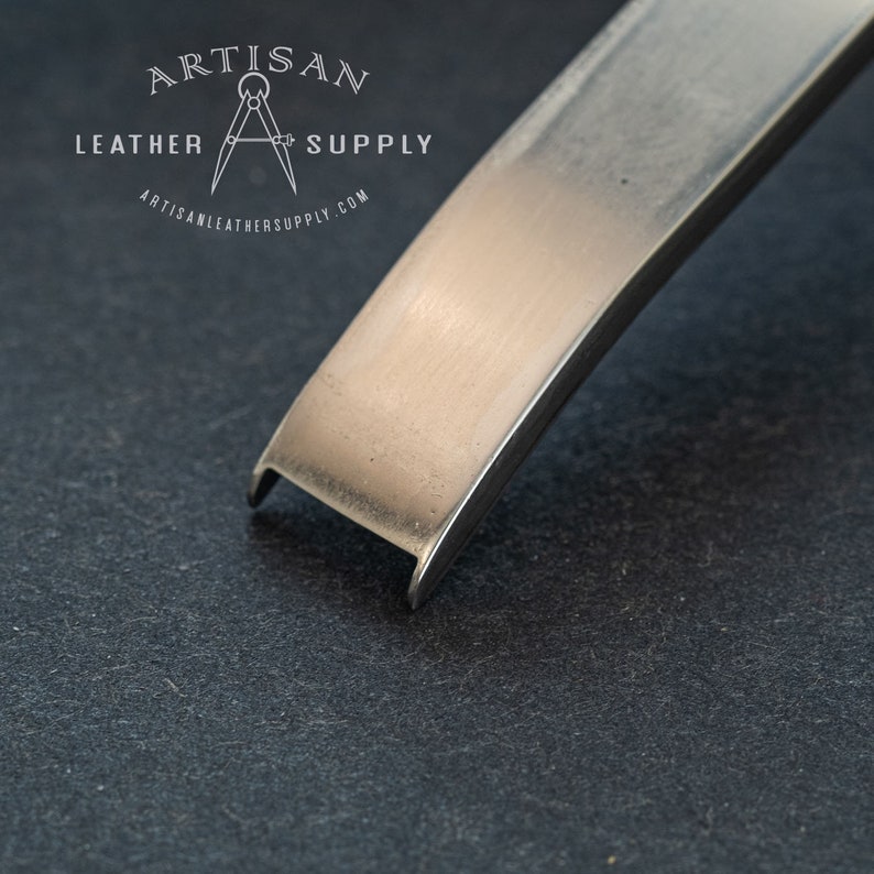 May include: Close-up of a silver metal edge beveler tool. The tool is designed for leather crafting and has a sharp, angled edge.